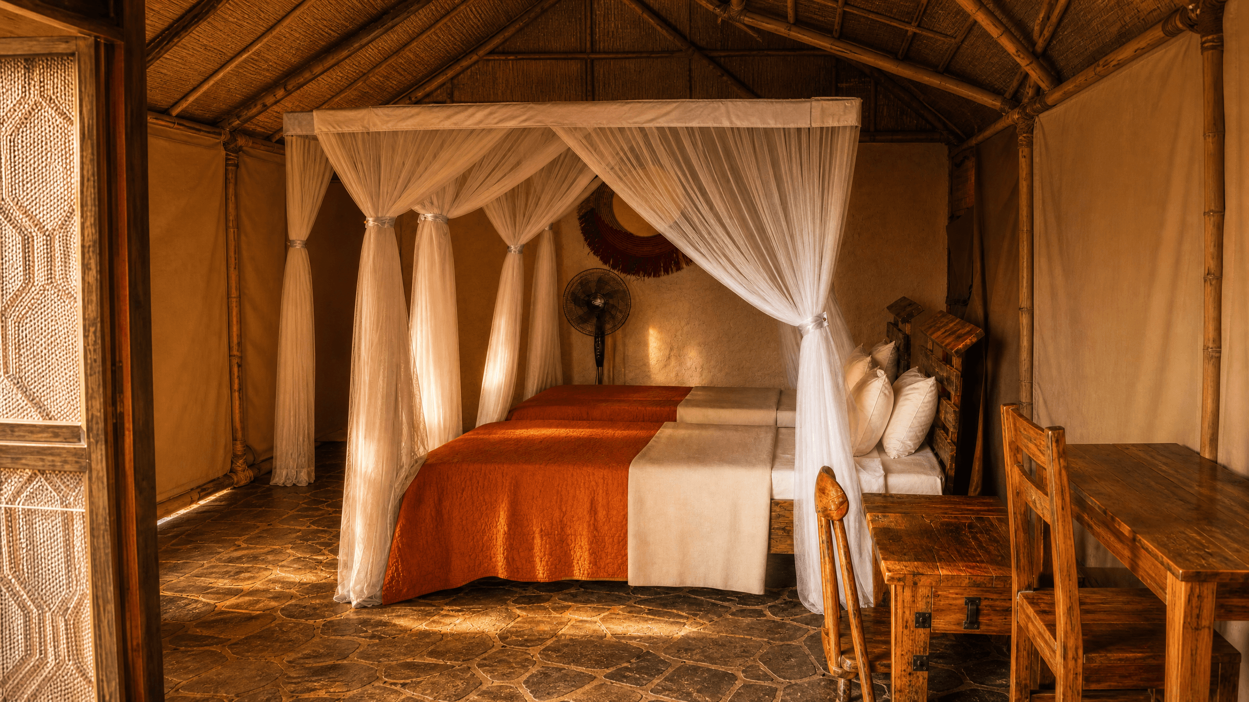 Deluxe Safari Tent bedroom at The Elephant Oasis — wide view showing twin beds with orange quilts, white mosquito net canopies, stone tile floors, canvas tent walls, bamboo ceiling structure, and wooden desk with chair, Murchison Falls Uganda