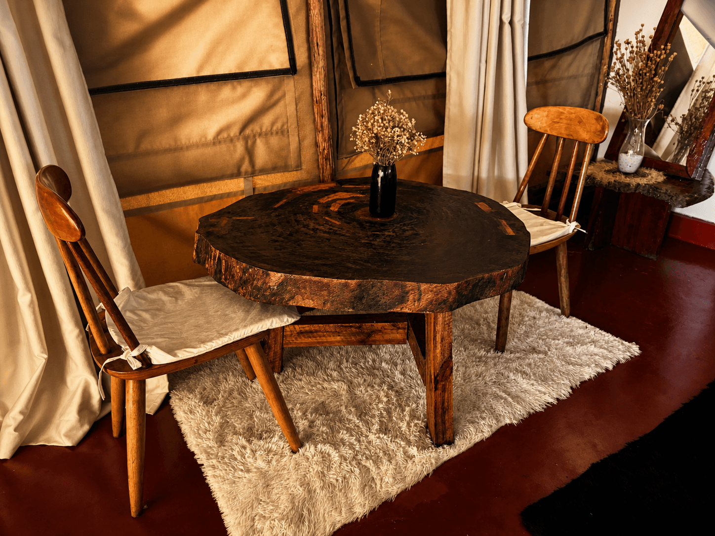 Luxury Safari Tent table and chairs at The Elephant Oasis — live-edge round cross-section table, two handmade wooden chairs with cushioned seats, dried flower arrangement, white shag rug, canvas tent walls, Murchison Falls Uganda