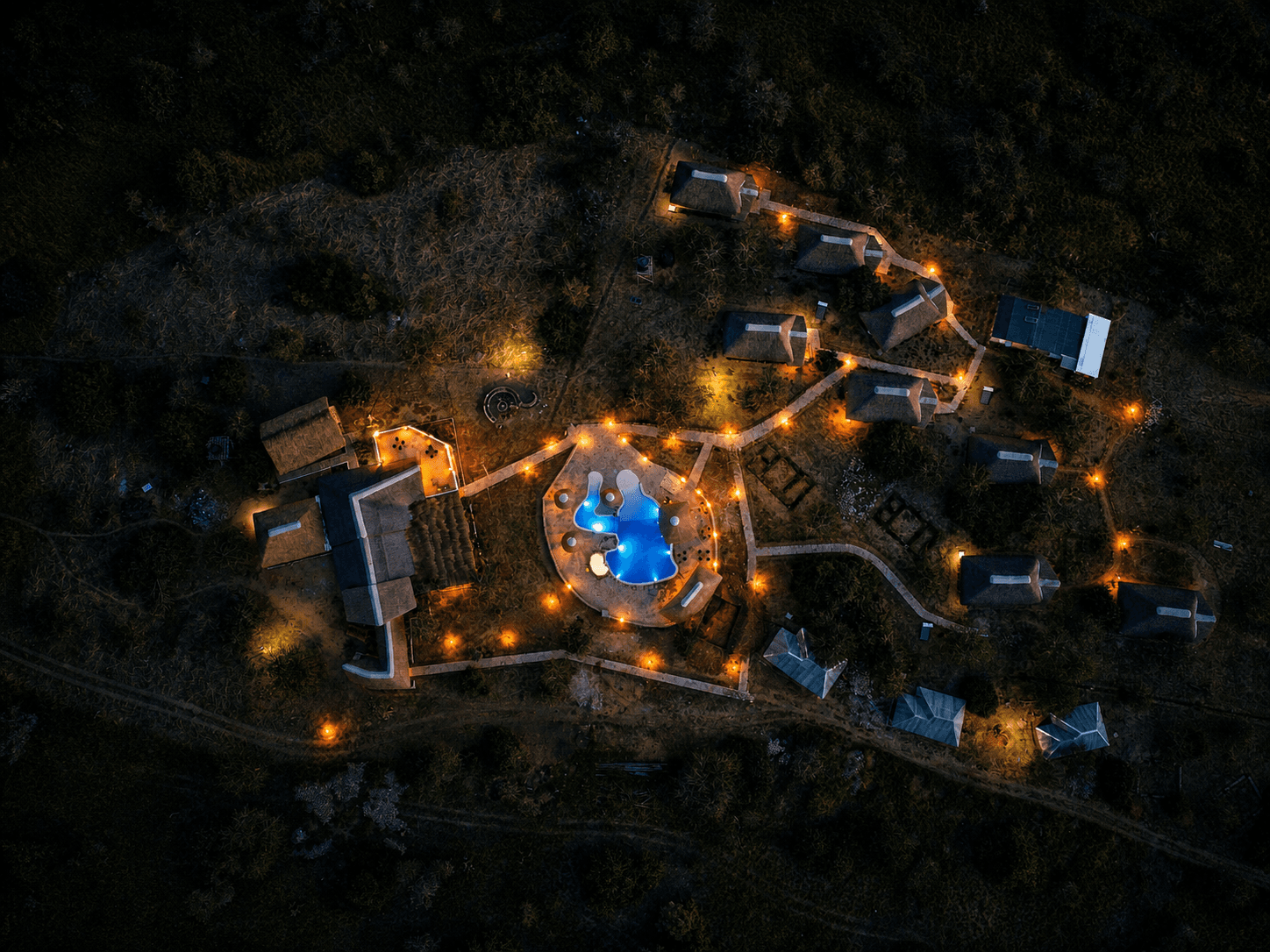 Night aerial of The Elephant Oasis — the elephant-shaped pool lit from within, tented accommodation, stone paths, and the Ugandan bush stretching to the horizon, Murchison Falls Uganda