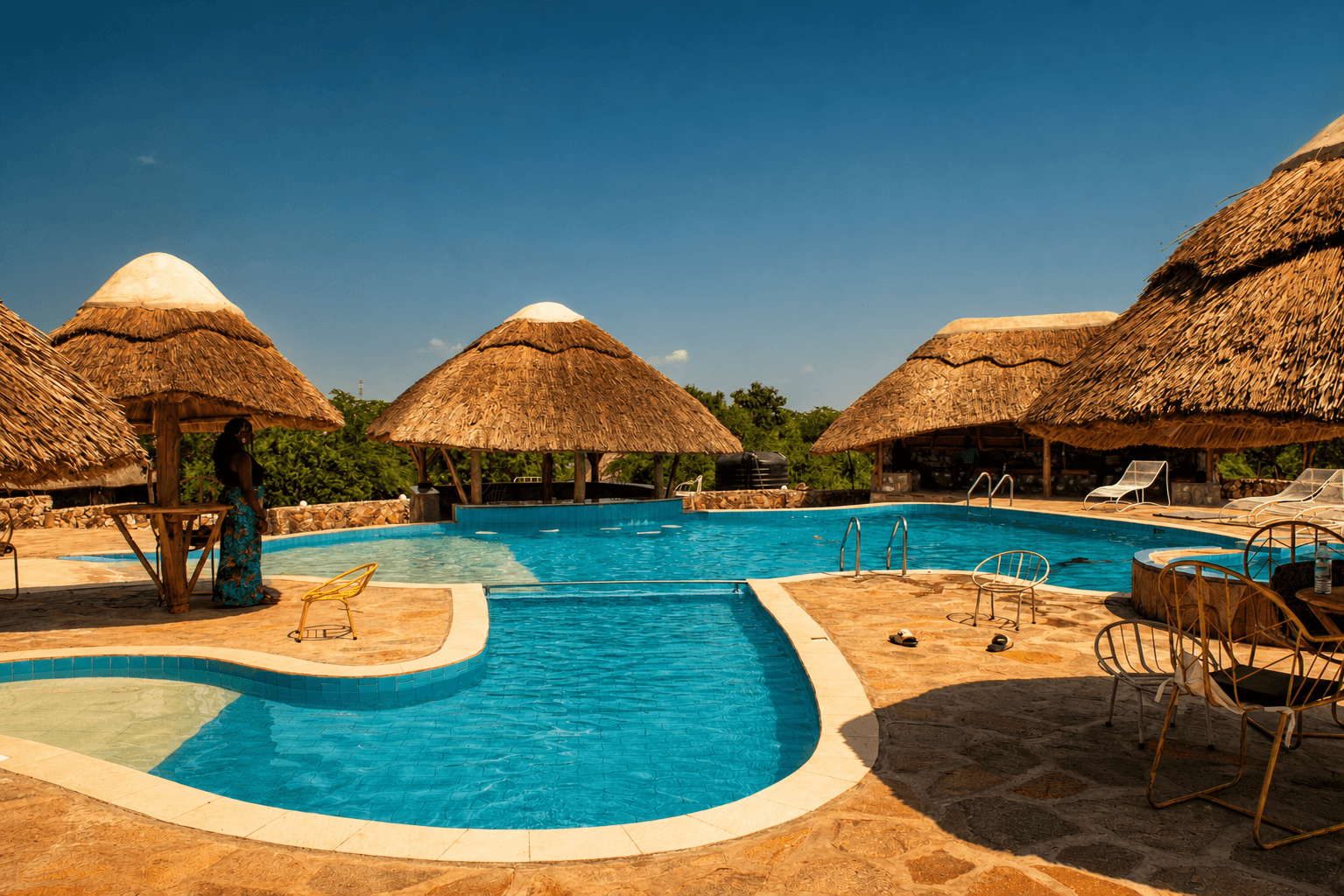 The elephant-shaped pool at The Elephant Oasis from ground level — turquoise water, thatched pavilions, and open sky, Murchison Falls Uganda